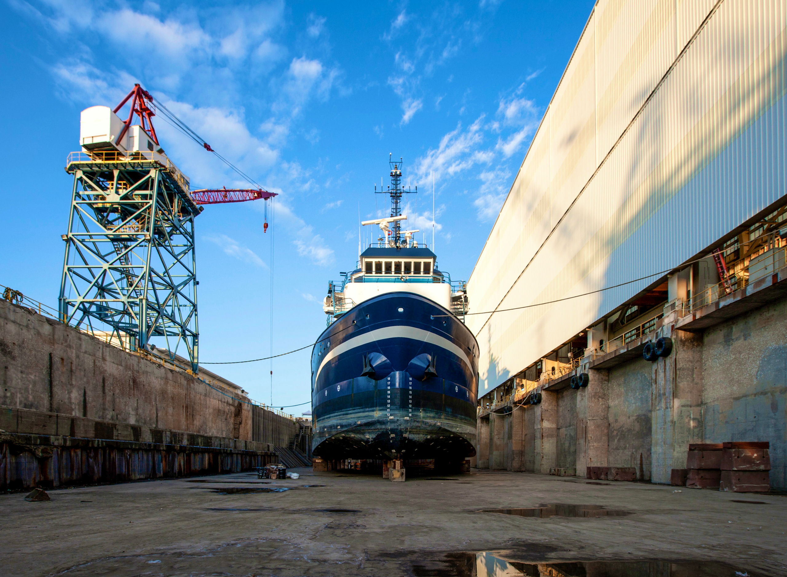 Large cargo ship positioned in a dry dock, surrounded by industrial structures including a crane and shipyard buildings under a clear blue sky.