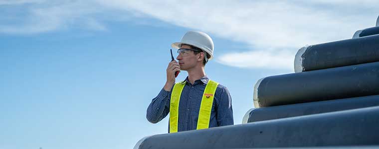 Worker wearing a hard hat and safety vest using a handheld radio near large industrial pipelines.